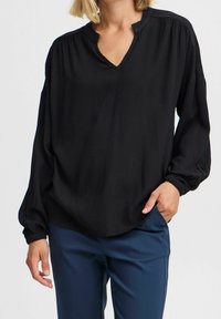 Kvinna som bär en lös svart texturerad långärmad blus och marinblå byxor, med handen i fickan, mot en enkel bakgrund.