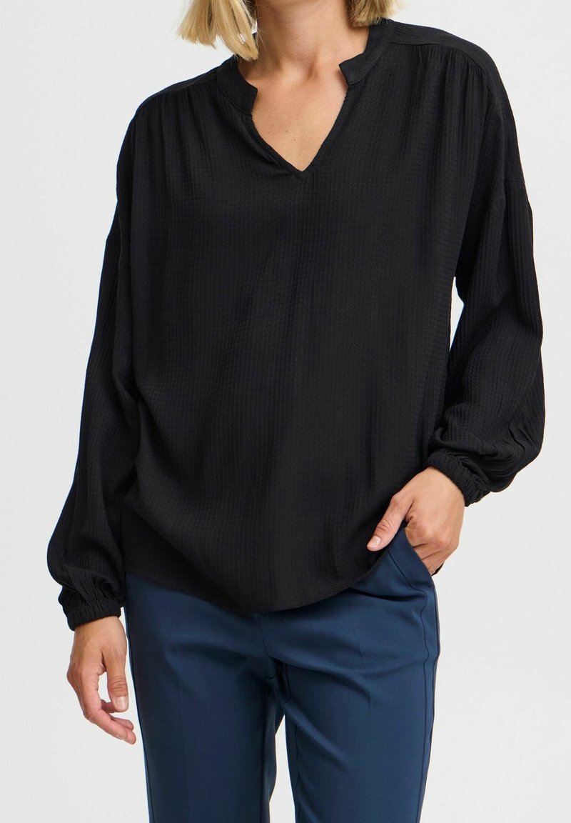 Kvinna som bär en lös svart texturerad långärmad blus och marinblå byxor, med handen i fickan, mot en enkel bakgrund.