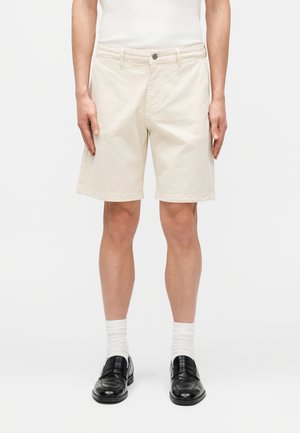 Muž v béžových šortkách po kolená, bielych ponožkách a čiernych kožených mokasínach stojaci na jednoduchom bielom pozadí.