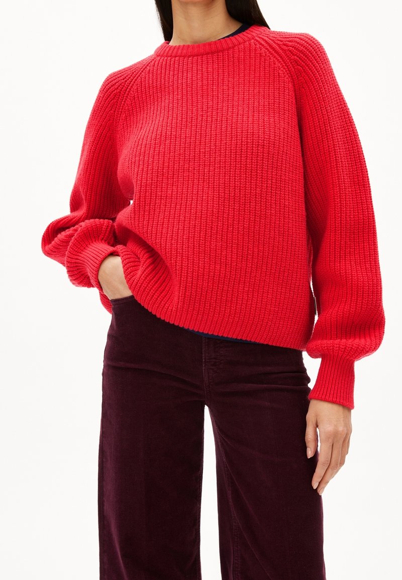 Femme portant un pull tricoté épais rouge vif à manches longues et un pantalon en velours côtelé bordeaux foncé, main dans la poche, sur un fond uni.
