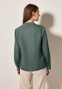 Camicia di lino verde con colletto, maniche lunghe e polsini abbottonati, mostrata di spalle su uno sfondo neutro.