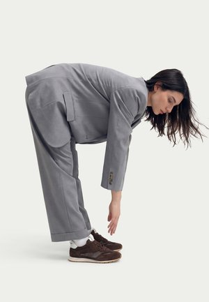 Femme en costume gris se penchant en avant pour toucher ses baskets marron sur fond blanc uni.