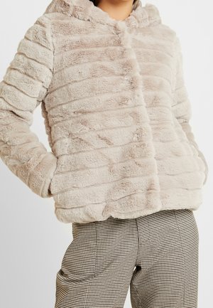 Beige Kunstfelljacke mit horizontalen, strukturierten Streifen, getragen mit schwarz-weiß karierten Hosen, Hände in den Jackentaschen.