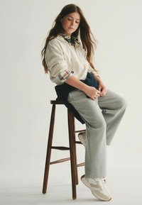 Licht beige zip-up hoodie, geruite shirtmanchetten, grijze sweatpants, marineblauwe gebreide riem, witte sneakers met beige accenten, zittend op een houten stoel.