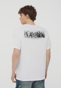 T-shirt in cotone bianco con una grafica in bianco e nero di un paesaggio urbano sulla schiena, con del testo sotto l'immagine in un carattere a contrasto.