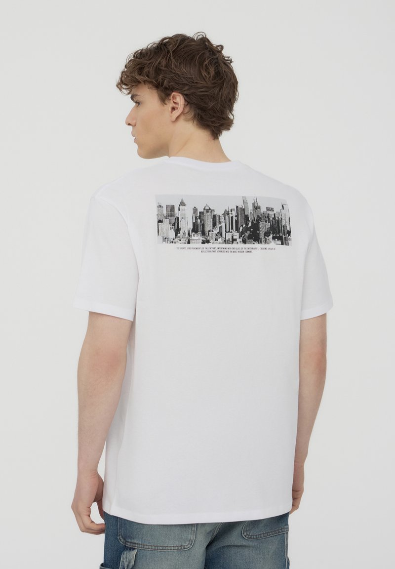 T-shirt in cotone bianco con una grafica in bianco e nero di un paesaggio urbano sulla schiena, con del testo sotto l'immagine in un carattere a contrasto.