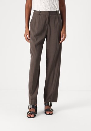 Personne portant un pantalon droit marron, des sandales à talons noires avec des boucles et un top sans manches blanc texturé, debout sur un sol blanc.