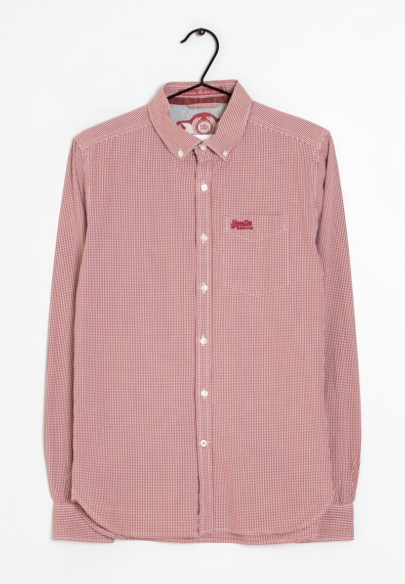 Chemise à manches longues à carreaux rouges et blancs avec un col boutonné et une poche. Présente un patch logo. Accrochée à un cintre noir.