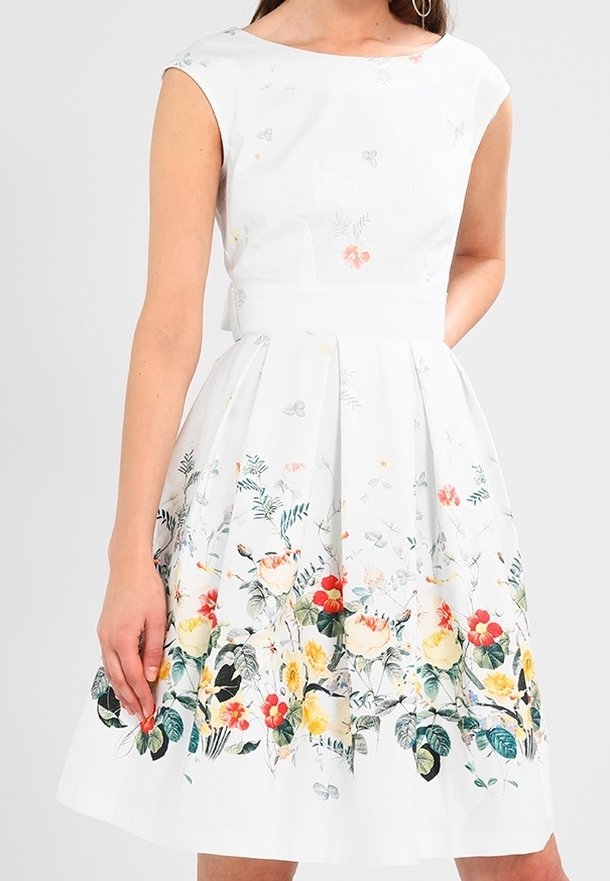 Femme portant une robe blanche sans manches avec un motif floral coloré autour de la jupe et de la taille.