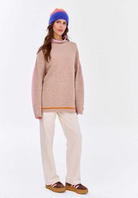 Hellbrauner gestrickter Pullover mit rosa Akzenten, hohem Kragen und oversized Ärmeln, kombiniert mit cremefarbenen weiten Hosen und braunen Sneakers.