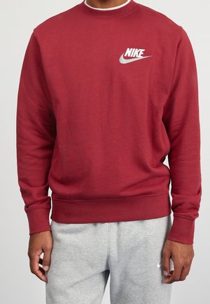 Mann trägt einen roten Nike-Sweatshirt mit weißem Logo auf der Brust und hellgraue Nike-Sweatpants, steht vor neutralem Hintergrund.