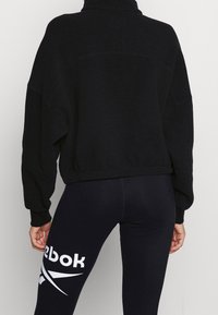 Černý fleece pullover s texturou, vysokým límcem a spadloumi rameny; v kombinaci s přiléhavými černými legínami s bílým logem.