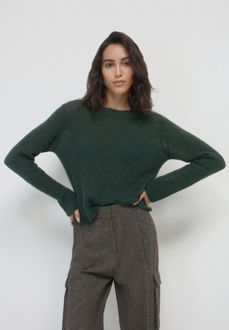 Dunkelgrüner, gestrickter Pullover mit lockerer Passform, langen Ärmeln und rundem Ausschnitt, kombiniert mit strukturierten braunen Hosen.