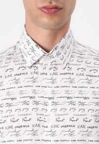 Hvit skjorte med knapp som har et gjentakende svart håndskrevet design, inkludert "Karl Lagerfeld" og andre signaturer, glatt tekstur.