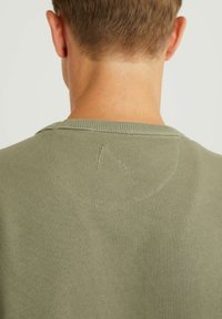 Camiseta de algodón color verde oliva, con escote redondo, costuras visibles y pequeño detalle bordado en la parte central de la espalda. Textura suave.
