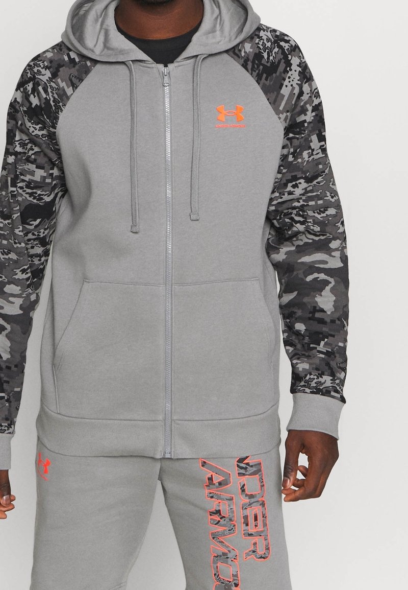Hombre con sudadera gris de Under Armour con mangas de camuflaje negro-gris y pantalones de chándal gris a juego con el logo y texto en naranja de Under Armour.