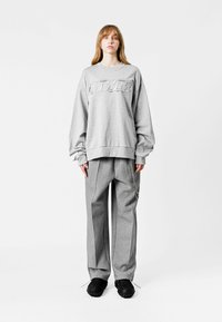 Sudadera gris con puños acanalados y un gran logo en relieve; combinada con pantalones grises holgados que tienen un detalle de cadena lateral y zapatos negros texturizados.