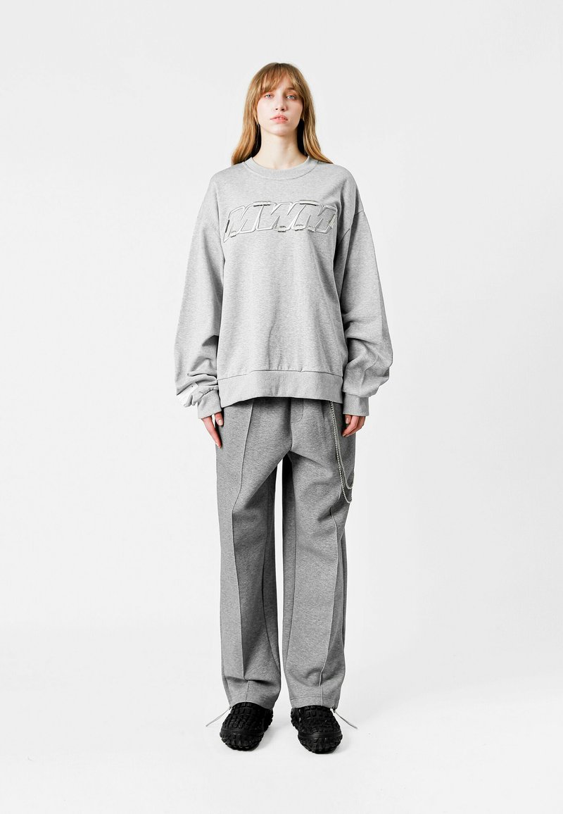 Sudadera gris con puños acanalados y un gran logo en relieve; combinada con pantalones grises holgados que tienen un detalle de cadena lateral y zapatos negros texturizados.