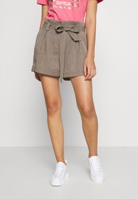 Lichtbruine, high-waisted shorts met een stoffen riem en steekzakken aan de voorkant, gecombineerd met een roze T-shirt met print en witte sneakers.