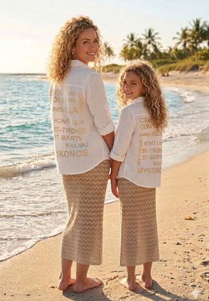 Zwei barfüßige Frauen, erwachsen und Kind, halten am Strand Händchen, tragen passende weiße Shirts mit Reisezielnamen und beige Röcke.