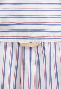 Gros plan d’un tissu blanc avec des rayures horizontales et verticales rouges et bleues, présentant une boucle en tissu beige cousue près du centre supérieur.