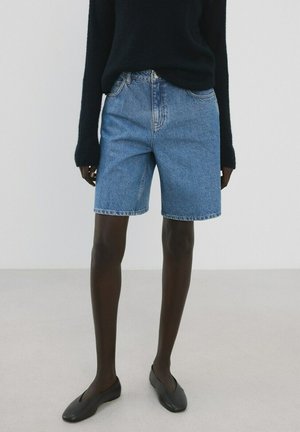 Osoba v modrých rifľových krátkych nohaviciach, čiernom dlhom rukáve a čiernych balerínach, stojaca pred jednoduchým svetlým pozadím.