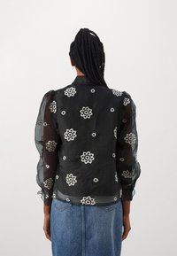 Blusa nera trasparente con ricami floreali, maniche a sbuffo e chiusura con bottoni sul retro. Presenta motivi floreali in bianco e argento a contrasto.