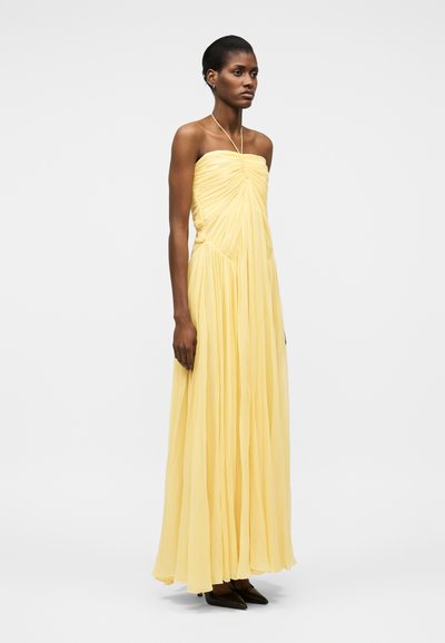 Vysoká žena v průsvitných, žlutých šatech bez ramínek s nařasením, stojící s uvolněnýma rukama, doplněná černými špičatými botami.