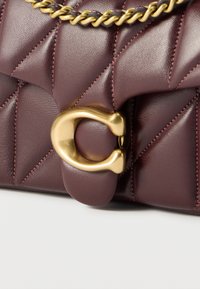 Σακίδιο από δέρμα σε σκούρο μωβ με καπιτονέ σχέδιο, με χρυσό κούμπωμα σε σχήμα C και αλυσίδα, σε κοντινή λήψη σε λευκή επιφάνεια.