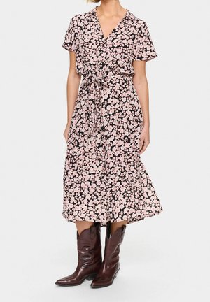 Mujer con un vestido midi rosa con estampado floral, mangas cortas y cintura atada, combinado con botas de cuero marrón oscuro hasta la mitad de la pantorrilla.
