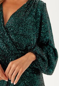 Abito verde smeraldo con una profonda scollatura a V, realizzato in tessuto ricoperto di paillettes, con maniche puff voluminose e dettagli in vita elastica.