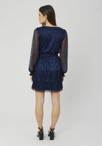 Robe bleu marine à volants avec des manches longues transparentes, en tissu texturé, taille élastique et ourlet à volants. Assortie à des chaussures à talons carrés noires.
