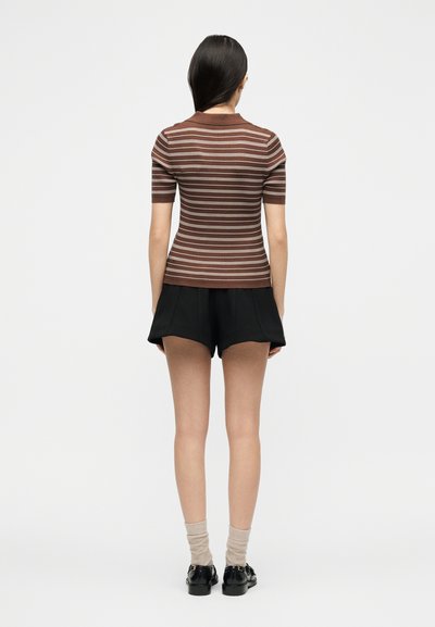 Femme aux longs cheveux foncés portant un haut à manches courtes rayé marron et blanc, un short noir, des chaussettes beiges et des chaussures noires, debout, vue de dos.