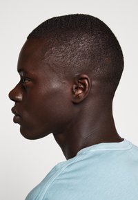 Vue de profil d'une personne à la peau foncée et aux cheveux courts et texturés, portant une chemise bleu clair à col rond.