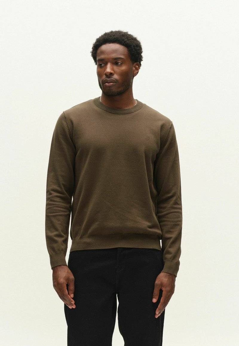 Homme aux cheveux courts et bouclés portant un pull marron à manches longues et un pantalon noir, debout devant un fond clair uni.
