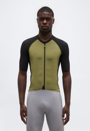 Άνδρας με εφαρμοστή κοντομάνικη ποδηλατική φανέλα σε ελιά και μαύρο χρώμα και γκρι επενδυμένο ποδηλατικό σορτς, στέκεται μπροστά σε απλό φόντο.