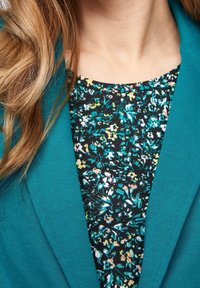 Blazer bleu sarcelle avec une finition texturée porté sur un haut noir à motif floral avec des accents verts, jaunes et roses, mettant en valeur un style superposé.