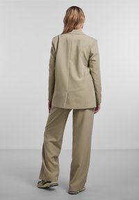 Femme debout de dos portant une veste de costume beige et un pantalon à jambes larges avec des baskets accentuées de néons sur un sol gris et un fond de mur blanc.