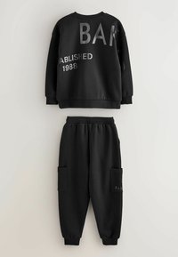 Czarny zestaw bluzy i dresów z miękkiego materiału. Bluza ma metaliczny napis "BAK" oraz "ZAŁOŻONA W 1988 R." Spodnie mają kieszenie cargo.
