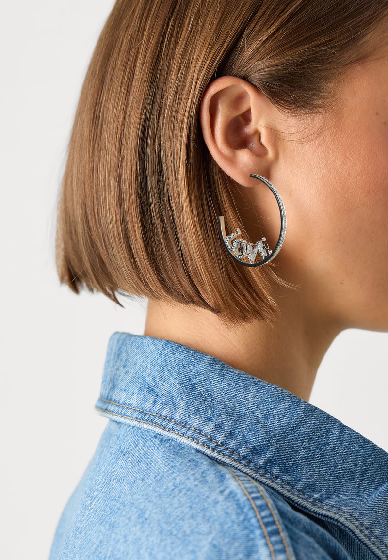 Boucle d'oreille en argent avec un design décoratif en forme de "love" orné d'accents transparents. Porté par une personne avec une coiffure bob courte, vêtue d'une veste en denim.