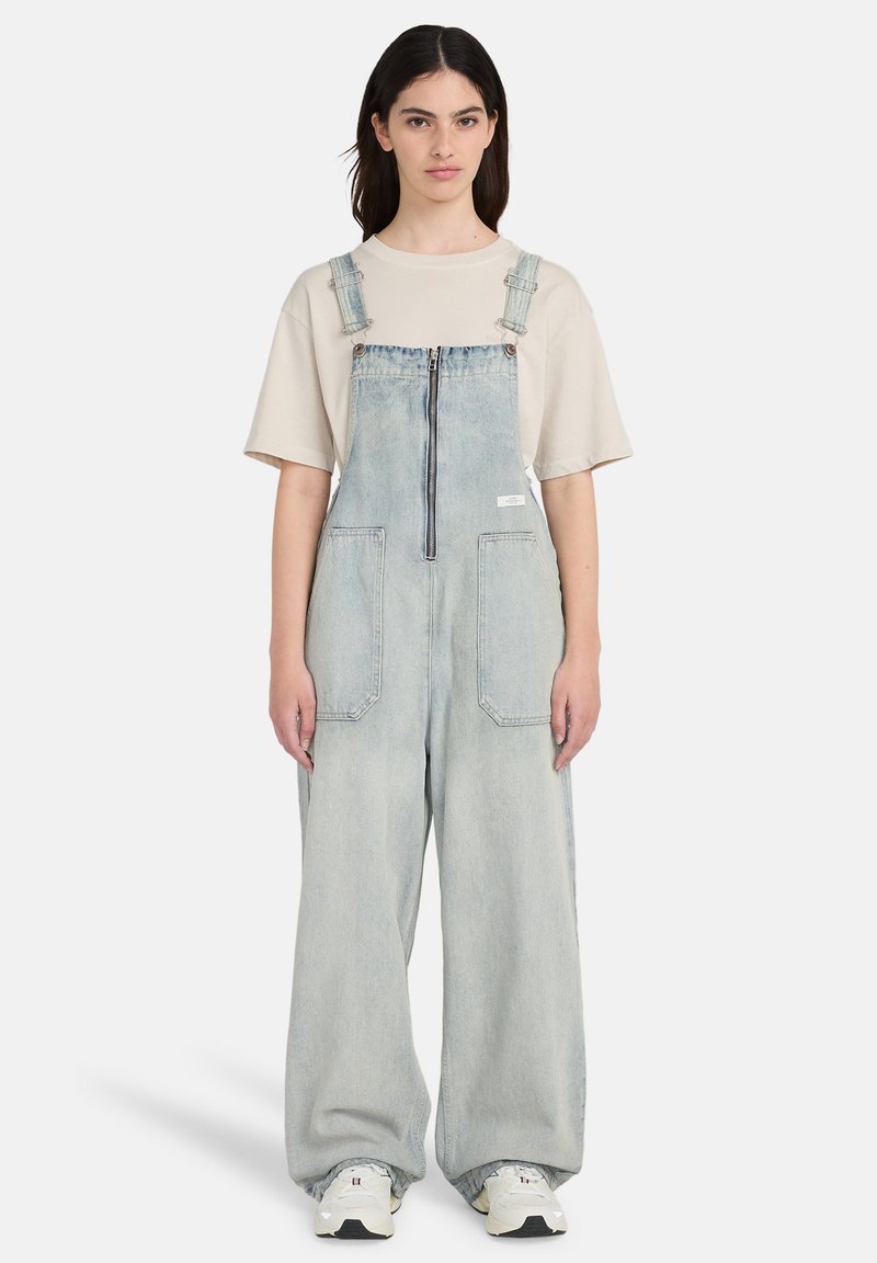 Salopette en denim bleu clair, dotée d'une fermeture éclair frontale, de bretelles réglables, de grandes poches avant et d'une coupe décontractée, associée à un t-shirt beige et des baskets.