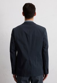 Granatowa marynarka z teksturowanej tkaniny, z wyraźnie zarysowanymi ramionami i pojedynczym rozcięciem z tyłu. Posiada wąskie klapy i zapinane mankiety.