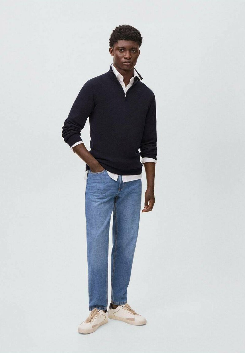 Mornarsko pulover s zip ovratnikom čez belo srajco, v kombinaciji z svetlo modrimi jeans hlačami in kremnimi supergami, stoji ob enobarvni ozadju.