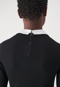 Robe noire côtelée avec un col blanc, dotée d'une fermeture éclair au dos. Le matériau semble lisse et ajusté, mettant en valeur une silhouette élégante.