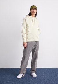 Krämfärgad hoodie med dragsnören och framficka, ihop med grå corduroybyxor. Vita sneakers med gula och lila detaljer. Olivgrön keps.