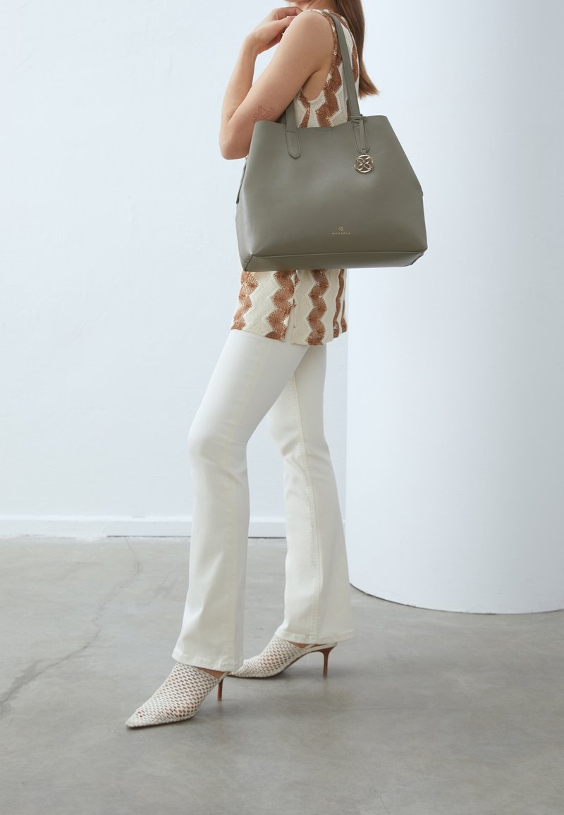 Olivgrön läderhandväska med två bärhandtag, prydd med en cirkulär metalldetalj och slät yta, tillsammans med en mönstrad topp och vita byxor.