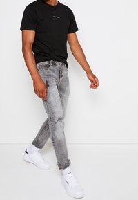 Svart bomullströja med liten vit logotyp, kombinerad med ljusgrå slitna jeans och vita sneakers, med upprullade ärmar och diskreta detaljer.