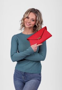 Červená psaníčko s texturovaným povrchem, ve tvaru obálky, s pozlaceným bleskem jako akcent, držené osobou v tyrkysovém žebrovaném svetru.