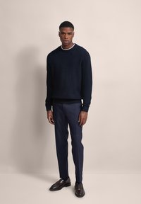 Marineblauer Strickpullover mit strukturiertem Muster, kombiniert mit maßgeschneiderten marinen Hosen und glänzenden braunen Schnürschuhen. Schlichter Hintergrund.
