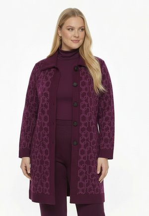 Vrouw met lang blond haar die een paarse jas met patroon draagt over een bijpassende coltrui en broek, staand tegen een witte achtergrond.
